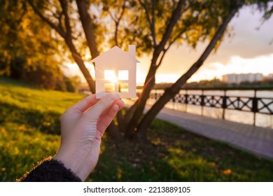 Hand Holds A Paper House Against The Background Of The Sunset Sky. The Sun Is Setting Over The Horizon. The Concept Of Your Own Home, Personal Space And Mortgage. High Quality Photo