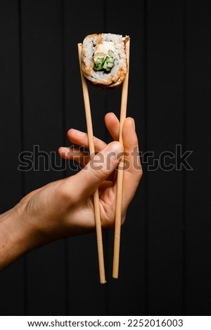 hand holds Philadelphia sushi roll with smoked eel, cucumber, avocado, cream cheese with bamboo chopsticks on dark background. Japanese food.