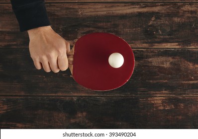 Hand Holds Professional Wooden Ping Pong Rocket With Ball On It. Red Attack Plus Pad On Rocket, All Above Old Vintage Table.