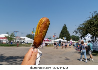 Hand holds up a pronto pup smothered in mustard at the fairgrounds in summer