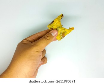 a hand holds a rectangular piece with a yellow base and inclusions against a white background. the piece has a textured appearance, suggesting a snack or food item