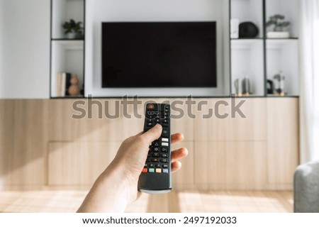 A hand holds a remote control in front of a flat screen TV mounted on a wooden entertainment center. Close up shot of woman press button, switch channel, watching movie at home. Copy space, mockup