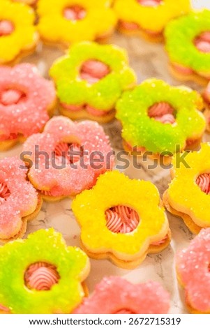 A hand holds a single Easter Cookie Sandwich with Raspberry Buttercream wrapped in clear cellophane. The cookie is flower-shaped with yellow sugar crystals and pink buttercream filling. Ideal for