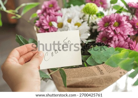 hand holds small note with text thank you on russian written on it, above a bouquet of pink white chrysanthemums wrapped in vintage newspaper. gratitude, kindness, and a warm gesture of appreciation.