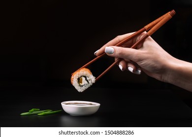 Hand holds sushi over bowl with soy sauce on black table.