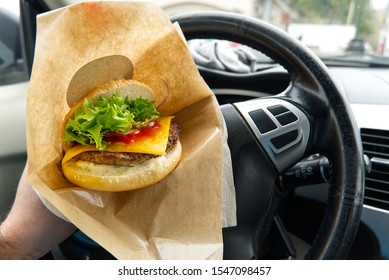 Hand Holds Tasty Hamburger Driving A Car. Closeup Photo