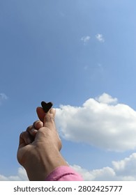 A hand holds a tiny, dark heart shaped object from soil. Against a vast, bright blue sky with soft white clouds over a field.