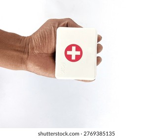 A hand holds a white rectangular box with a red cross emblem on the front against a plain white background.