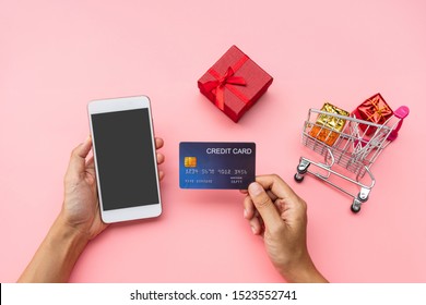 Hand Holing Credit Card And Mobile Phone, Shopping Cart With Gift Boxes On Pink Background. Shopping Online, Modern, Convenient Lifestyle, New Normal Concept. Copy Space, Top View