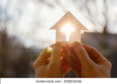 Hand with a house from paper and a cross on a background of sunrise. - Powered by Shutterstock