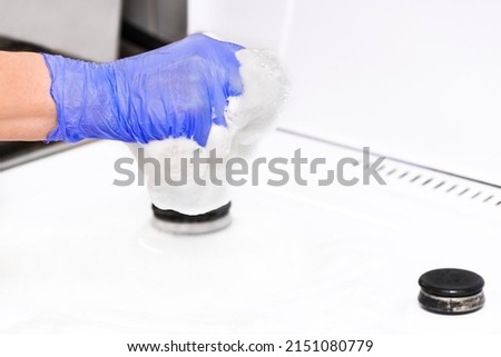The hand of a housewife woman in a latex household glove squeezes foam from a sponge to wash a gas stove in the kitchen, close-up.