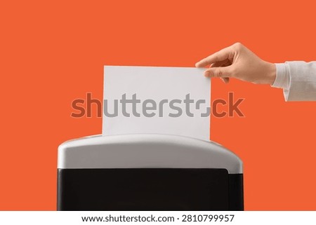 Hand inserting a blank white paper sheet into a document shredder for destruction and data security on an orange background