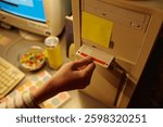 Hand inserting floppy disk into old desktop computer tower with yellow note and candies nearby in soft light. Retro setup featuring vintage technology and personal touch