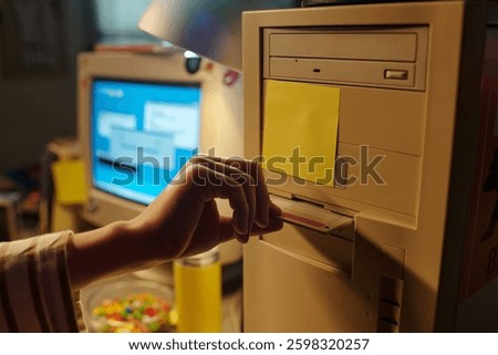 Hand inserting floppy disk into vintage computer with background showing another computer monitor displaying a blue screen. Scene capturing nostalgia of retro technology
