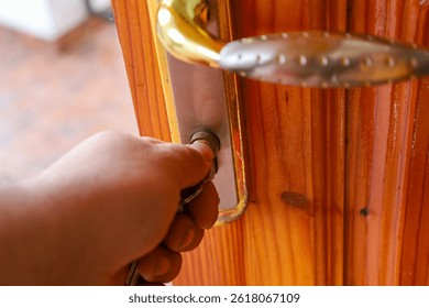 A hand inserts a key into the lock of a wooden door with a golden handle and a metal plate. The silver-colored key is partially inserted into the lock cylinder. The door has a pronounced wood texture.