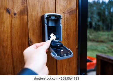 A hand inserts a key into an open wall mounted lockbox on a wooden surface. An orange vehicle is partially visible, with trees in the distance.