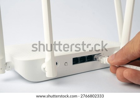 A hand inserts a network cable into a port on a white router. The router has multiple antennas and ethernet ports.