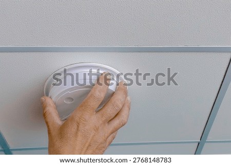 hand installing or checking smoke detector on ceiling tile indoors. Fire safety maintenance, home protection and alarm system concept.