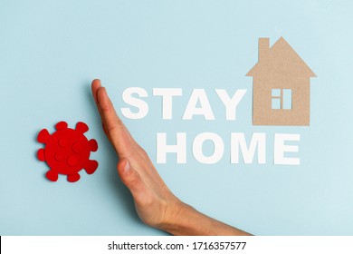 Hand Keeping Away Red Coronavirus Shape From Cardboard House. Message Next To It States Stay Home On A Blue Surface. Self Isolation Concept. Protect Your Home Concept.