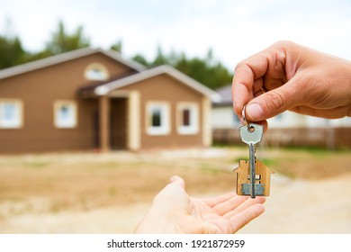 Hand with a key and a wooden key ring-house. Background of fence and cottage. Building, project, moving to a new home, mortgage, rent and purchase real estate. To open the door. Copy space
