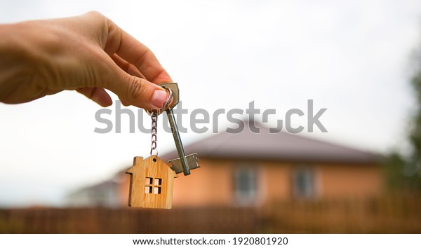 Hand with a key and a wooden key ring-house.\
Background of fence and cottage. Building, project, moving to a new\
home, mortgage, rent and purchase real estate. To open the door.\
Copy space