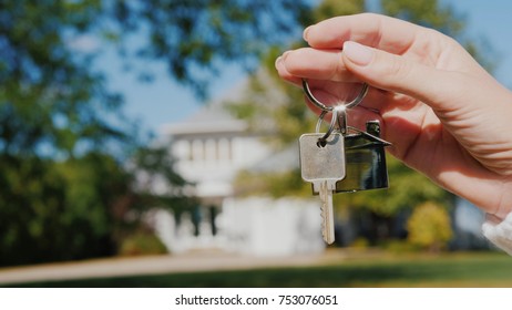 A hand with a keychain in the shape of a small house and keys. Against the background of a typical house in the usa suburban style