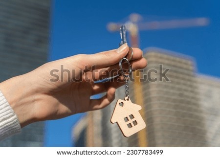 Hand with keychain in shape of small house and keys. Against backdrop of typical American country-style house. Construction, sale or purchase of apartments. Developer, realtor or real estate agency.