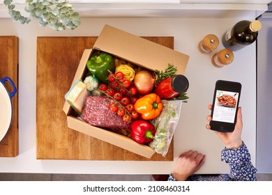 Hand In Kitchen Holding Phone With Recipe On Screen For Online Food Recipe Kit Delivered To Home