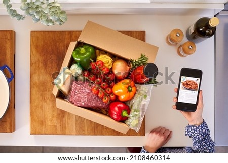 Hand In Kitchen Holding Phone With Recipe On Screen For Online Food Recipe Kit Delivered To Home