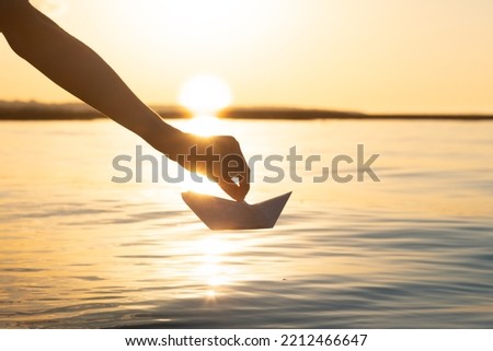 the hand launches the paper ship into the water. Beautiful summer sunset on the river and origami paper boat