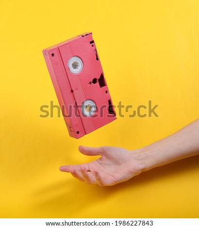Hand and Levitating pink video cassette on yellow background. Minimalistic still life. Concept art. Video game