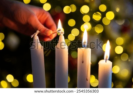Hand lighting third candle with a match on Advent wreath with four pillar candles. Christmas tree with twinkling lights
