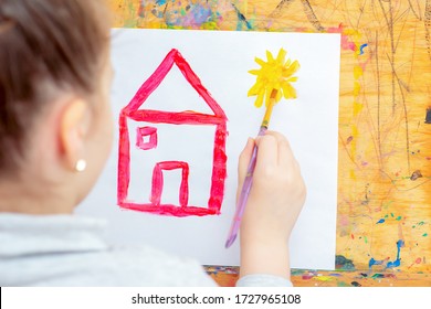 Hand of Little girl drawing yellow sun and red house with brush on easel at home. Children's creativity.