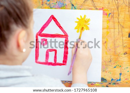 Hand of Little girl drawing yellow sun and red house with brush on easel at home. Children's creativity.