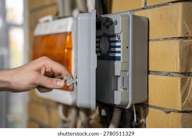 Hand locking outdoor irrigation controller cabinet with a key, side view of automatic watering control box mounted on brick wall.