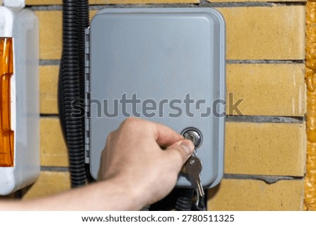 Hand locking outdoor irrigation controller cabinet with a key, side view of automatic watering control box mounted on brick wall.