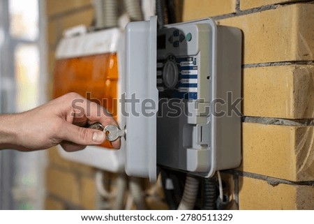 Hand locking outdoor irrigation controller cabinet with a key, side view of automatic watering control box mounted on brick wall.