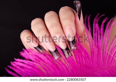 Hand with long artificial french manicured nails colored with black nail polish with glitter and gemstones decoration and pink feather. Black background.