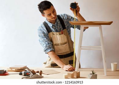 Hand Made Furniture Concept - Caucasian Male Woodworker In Apron Measuring Wooden Stool With Flexible Ruler, Custom Made Furniture And Handicraftship Concept