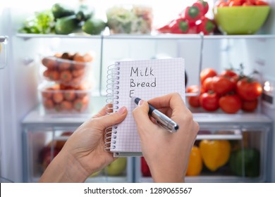 Hand Making List Of Food On Spiral Notepad In Front Of An Open Refrigerator