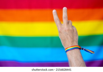 Hand Making The Symbol Of Peace With A Bracelet In Support Of The Lgtbi Collective; With The Lgtbi Flag In The Background