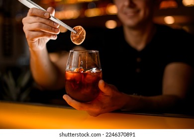 hand of male barman neatly holds glass with cold cocktail and garnishes it with slice of dry orange using tweezers