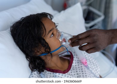 Hand Of Male Doctor Putting Oxygen Mask Ventilator On Sick Mixed Race Girl In Hospital Bed. Medicine, Health And Healthcare Services During Coronavirus Covid 19 Pandemic.