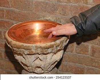 hand of man and an ancient holy water font inside the Church in a copper vase