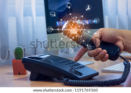 Hand of man holding telephone handset and pressing button on keypad, office desk with cactus pot, computer background, ip telephony services concept with flying icon and connectivity