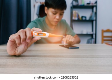 In The Hand Of The Man Holding A Thermometer To Measure The Temperature And Look At The Average, Check The Temperature Of Sickness Through The Phone On The Table. In The Room By Himself