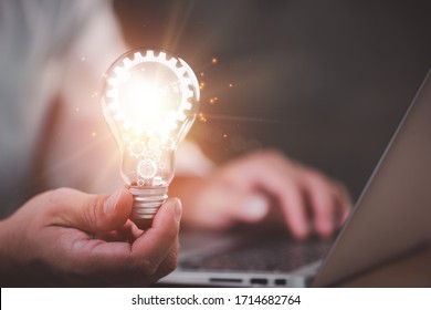 The hand of a man with a light bulb And there is a gear icon in it And he is using a notebook computer. The idea of ​​inspiration from online technology.innovation idea concept. - Shutterstock ID 1714682764