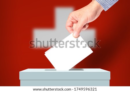 The hand of man putting his vote in the ballot box with flag Switzerland on background. Election in Switzerland