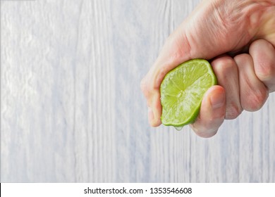 The hand of a man squeezes the juice from a slice of lime half. Juicy drop hanging down. Light wooden background.