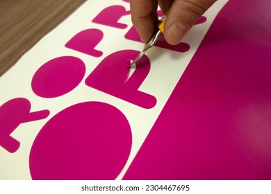 The hand of a man with a stationery knife, selects a vinyl film after cutting on a plotter.Sampling of the film in the production of avtovinil. Cutting a pink sticker. - Shutterstock ID 2304467695